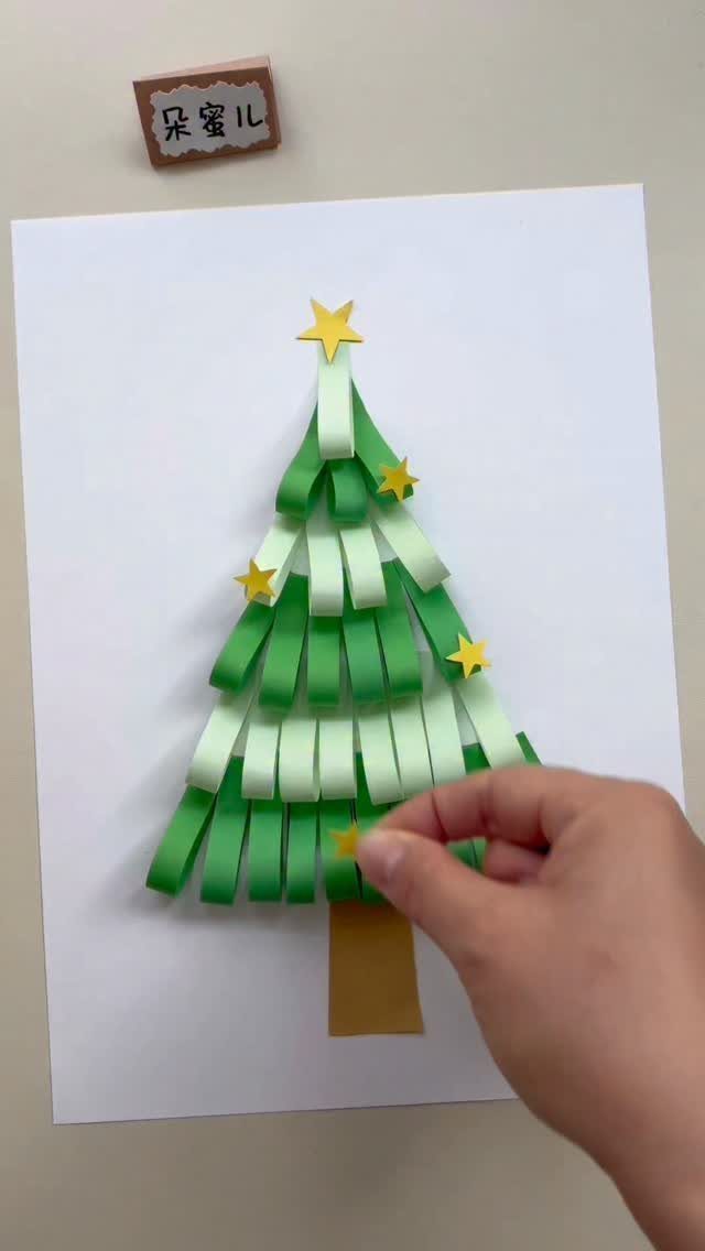 A hand placing a yellow star onto a green paper strip Christmas tree craft.