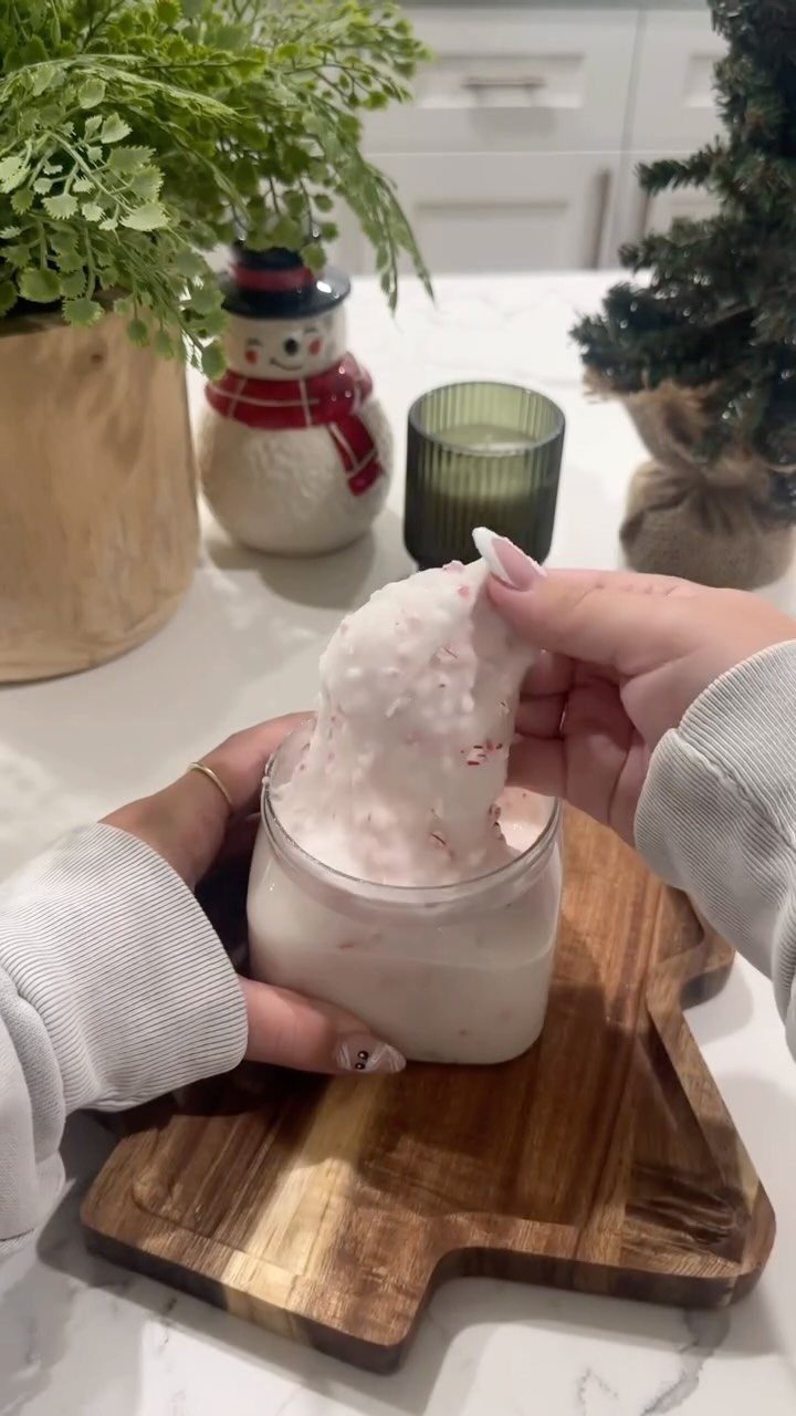 Hands scooping pink and white peppermint sugar scrub from a clear glass jar on a wooden board.