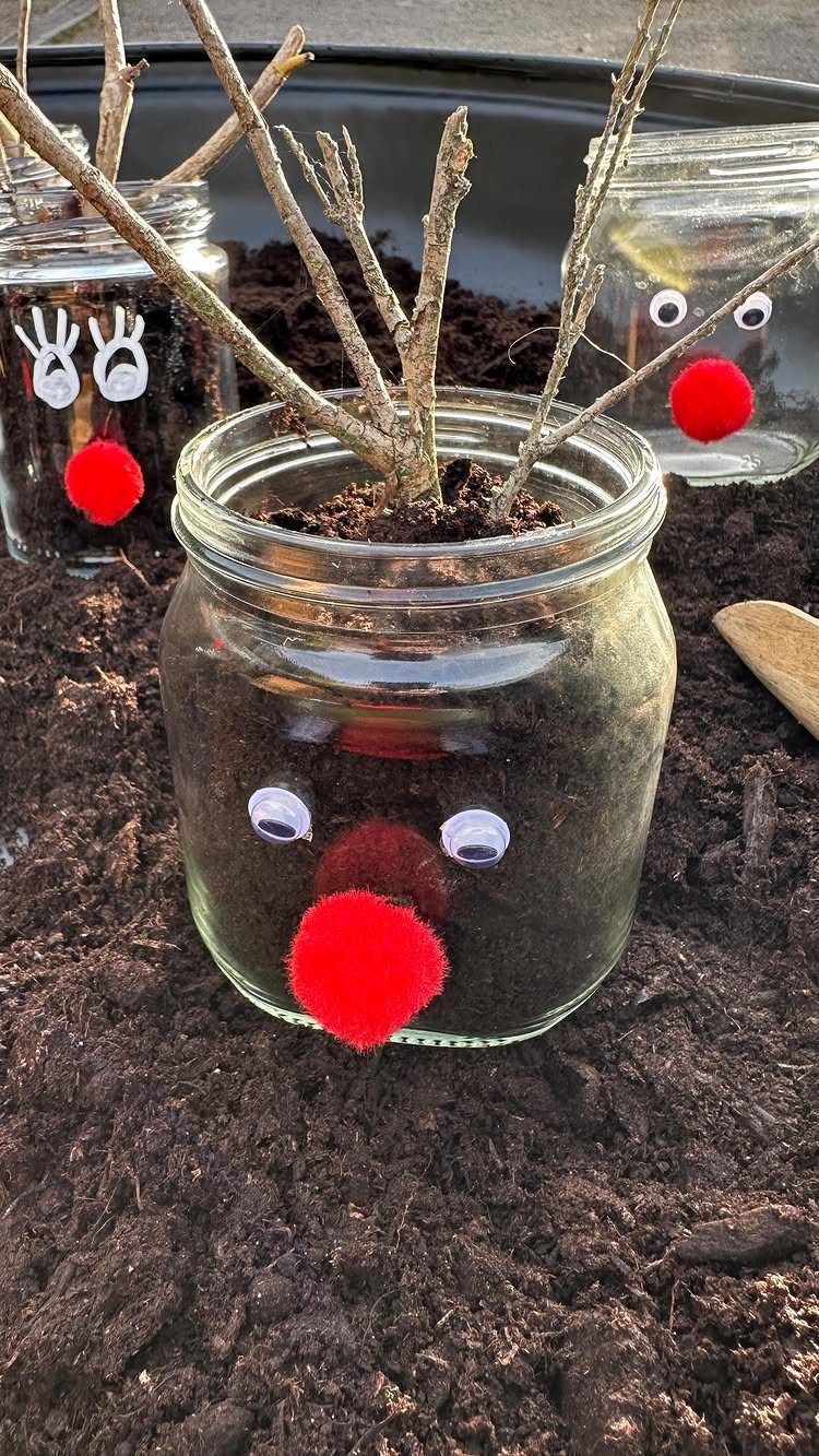 Three mason jars decorated as reindeer with red pom-pom noses and twig antlers in soil.