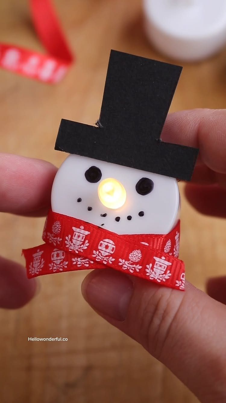 A person holding a small snowman ornament made from a white tea light candle with a red scarf.