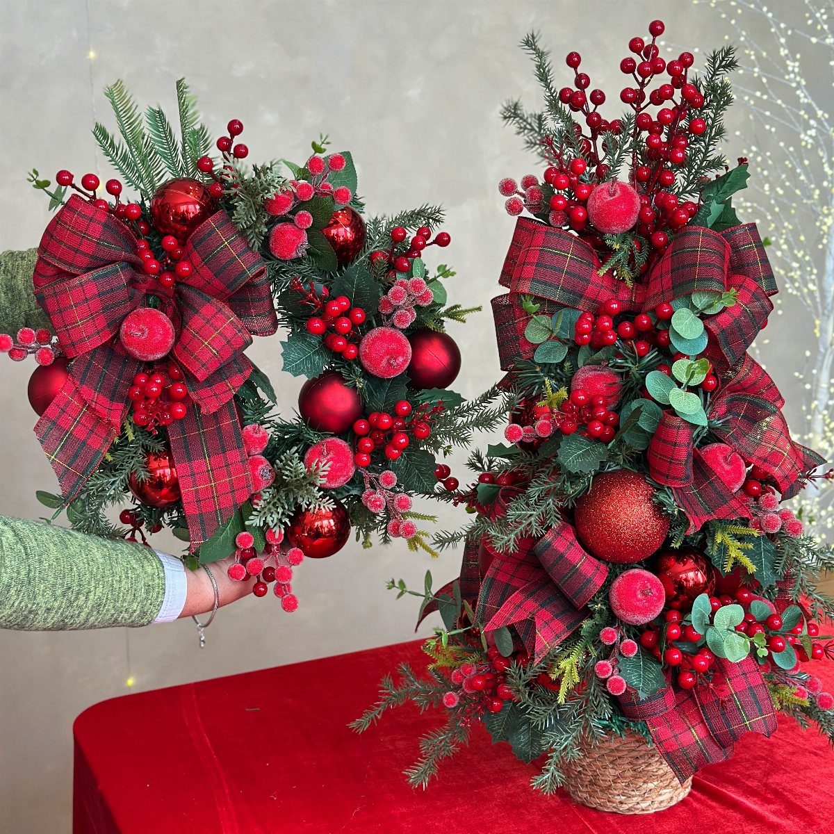 A person holding a festive Christmas wreath with red plaid bows and red berries.