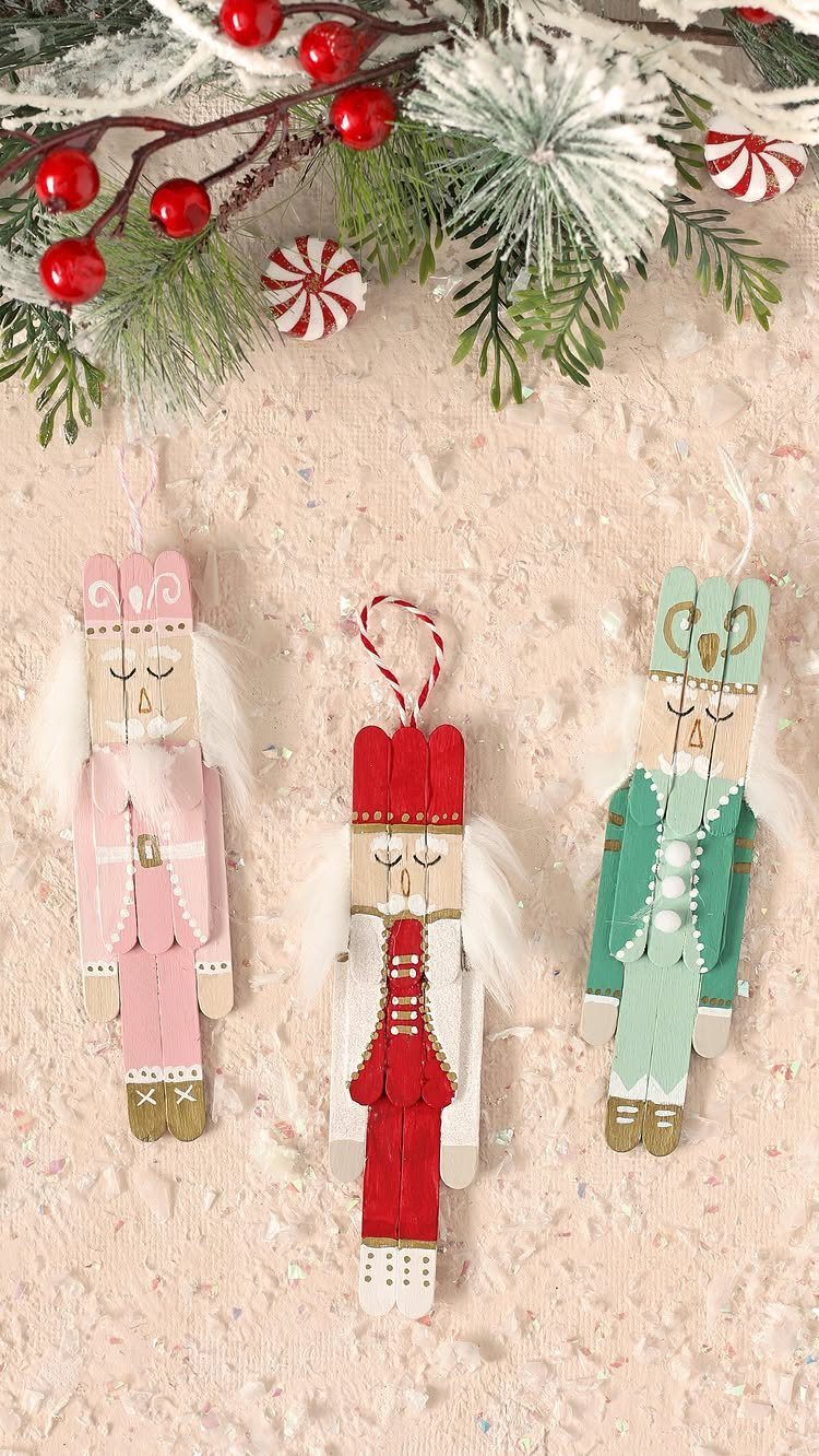 Three painted craft stick ornaments with white feather wings hanging against a pink background.