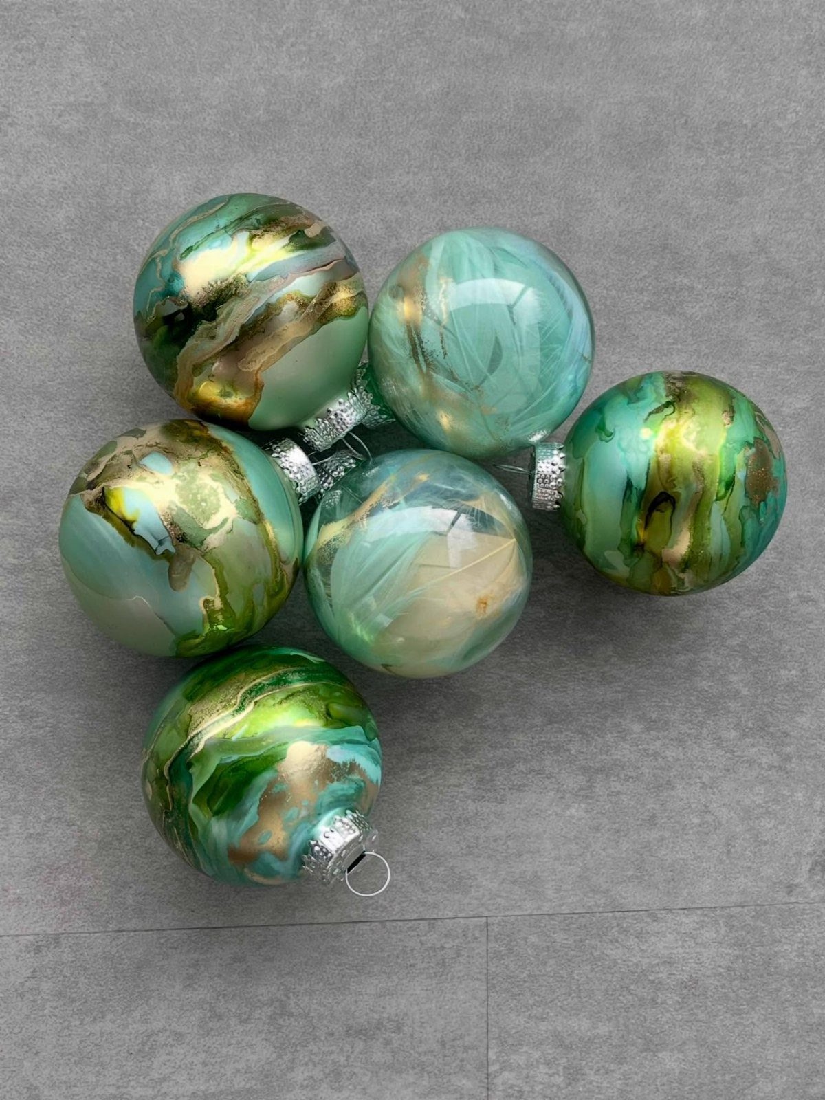Six glass Christmas ornaments featuring swirling green and gold alcohol ink patterns on a grey background.