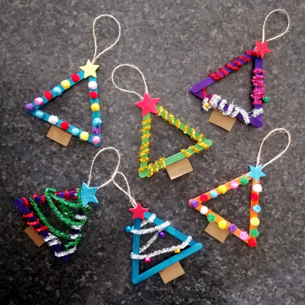 Six colorful craft stick Christmas tree ornaments with glitter and pom-poms on a dark surface.