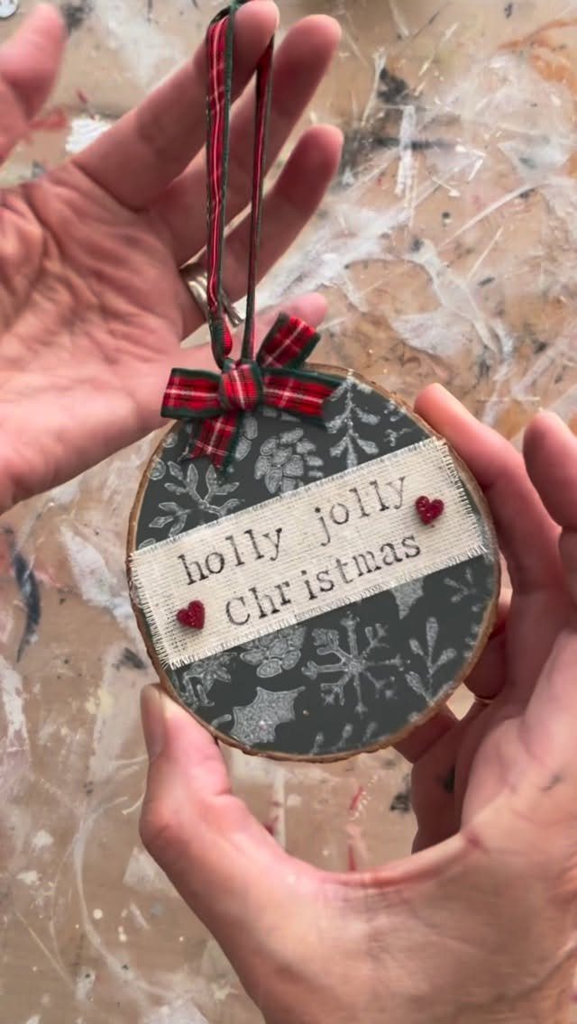 Hands holding a dark green wood slice ornament with white stenciled snowflakes and festive text.
