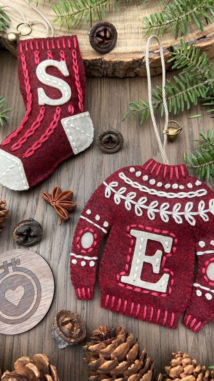 Red felt Christmas ornaments shaped like a stocking and a sweater resting on a wooden surface with pine branches.
