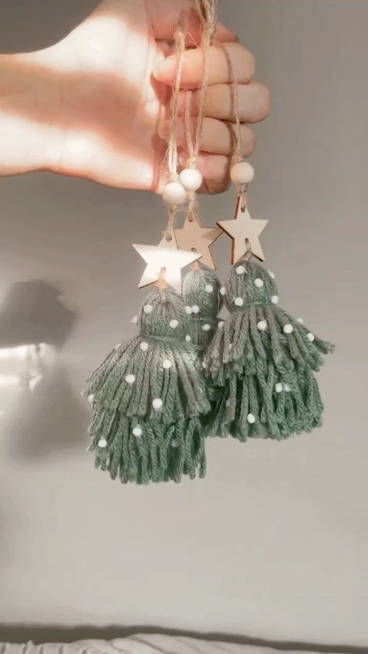 A hand holding three sage green yarn tassel ornaments with wooden star charms.