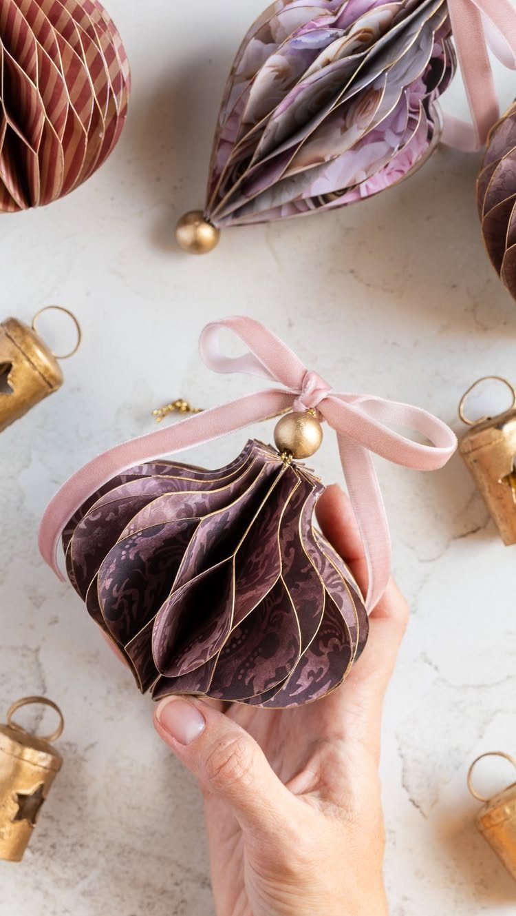 26 Handmade Ornaments: DIY Projects from Paper to Pinecones 17 A hand holding a purple patterned paper honeycomb ball ornament with a pink ribbon.
