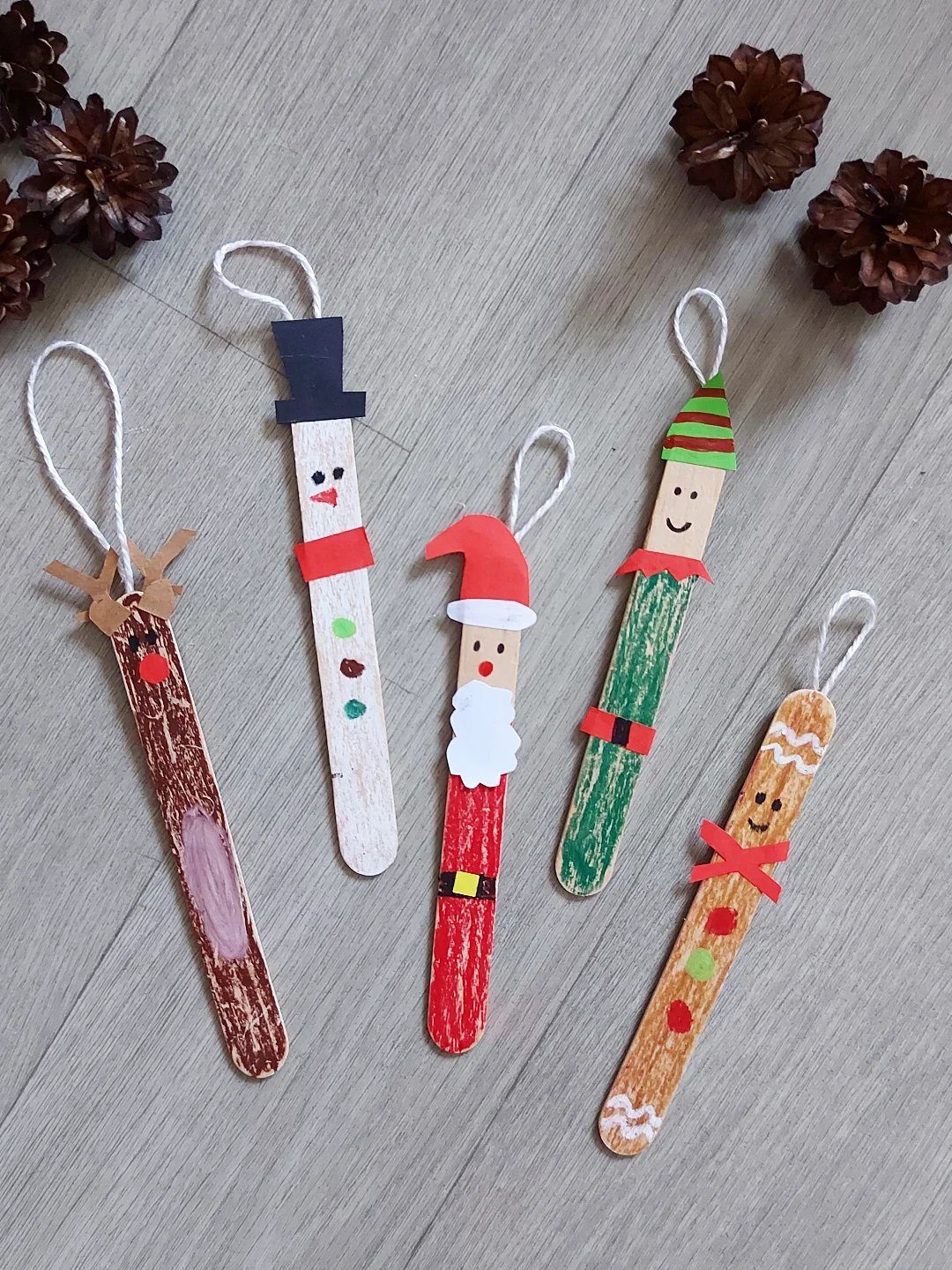 26 Handmade Ornaments: DIY Projects from Paper to Pinecones 11 Five decorated popsicle stick Christmas ornaments arranged on a light wooden surface with pinecones.