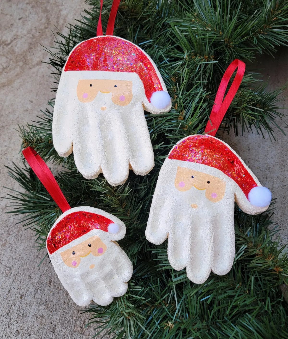 26 Handmade Ornaments: DIY Projects from Paper to Pinecones 5 Three Santa Claus handprint ornaments hanging from green pine branches with red ribbons.