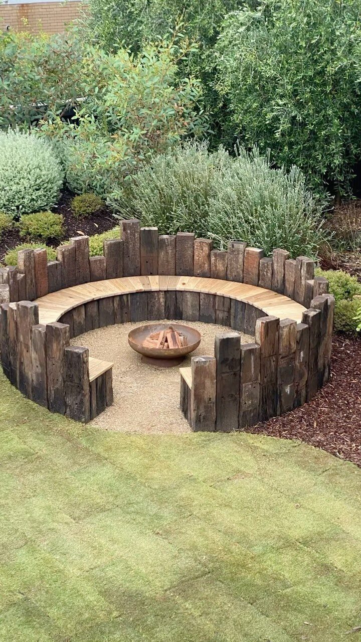 11 Inexpensive Landscaping Ideas for a Better Yard 3 A curved wooden log bench surrounding a small fire pit in a green garden.
