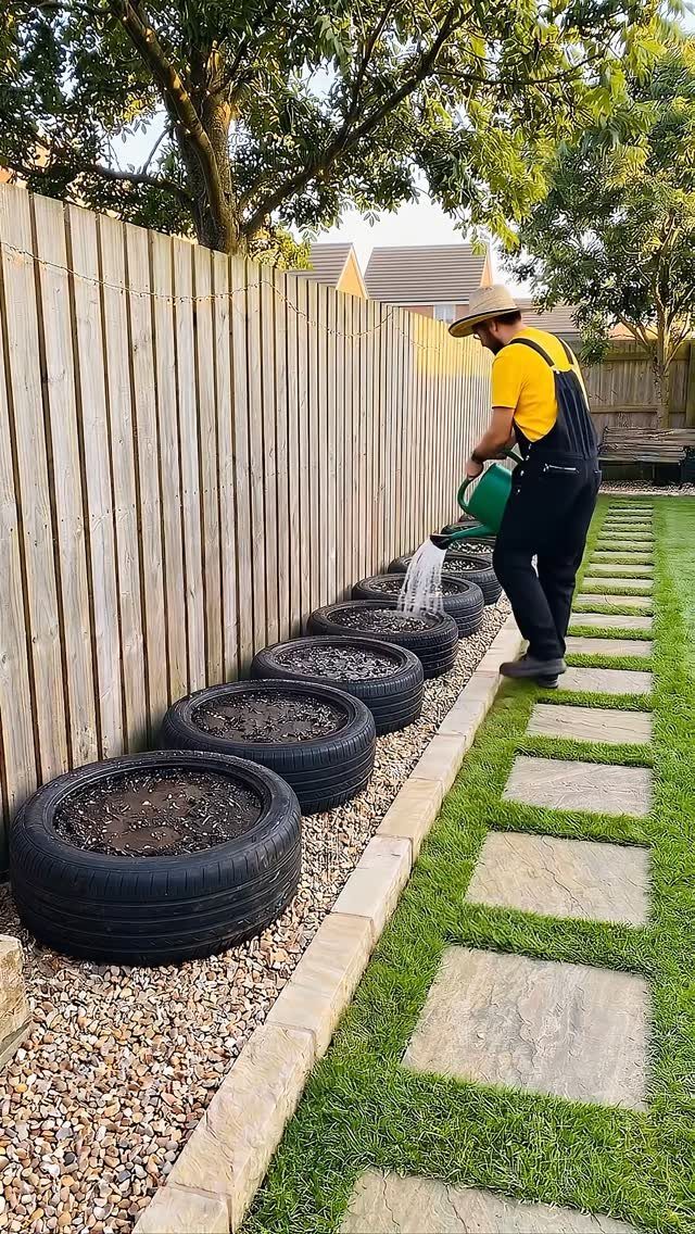 11 Inexpensive Landscaping Ideas for a Better Yard 1 A man watering plants growing inside black tires used as garden planters along a fence.
