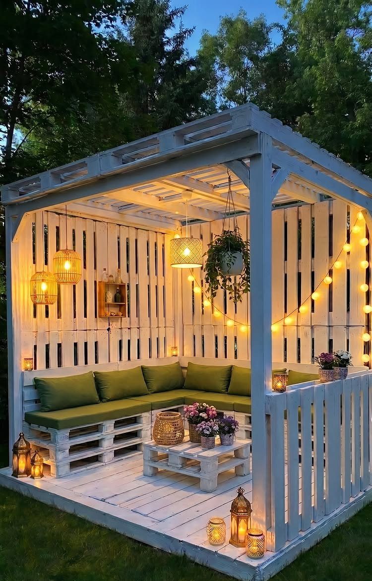 11 Inexpensive Landscaping Ideas for a Better Yard 11 White pallet gazebo with green cushions, string lights, and lanterns in a lush garden setting.