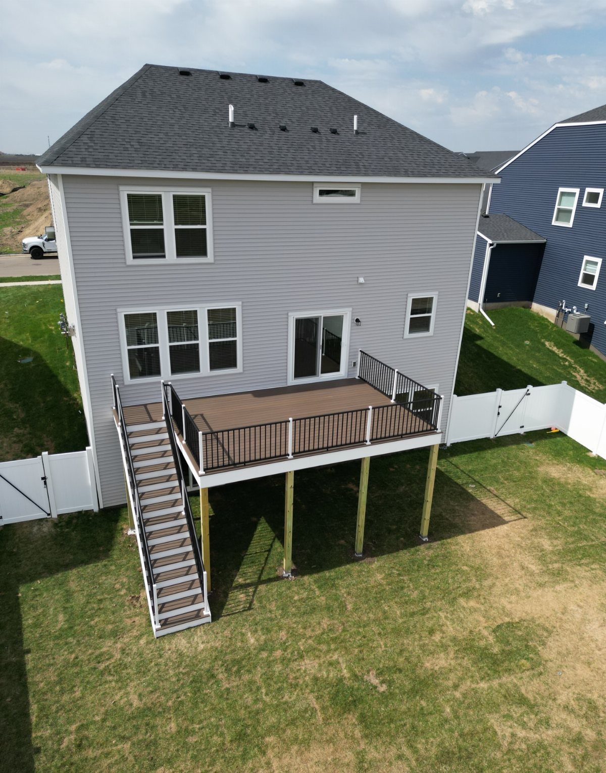 An elevated wooden deck with dark railings attached to the back of a grey two-story house.