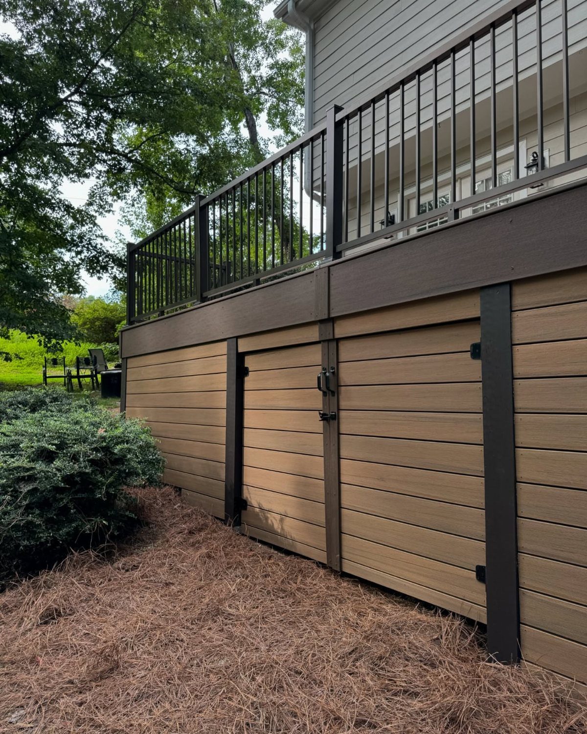 A raised wooden deck featuring light brown horizontal skirting and dark brown metal railings near green trees.
