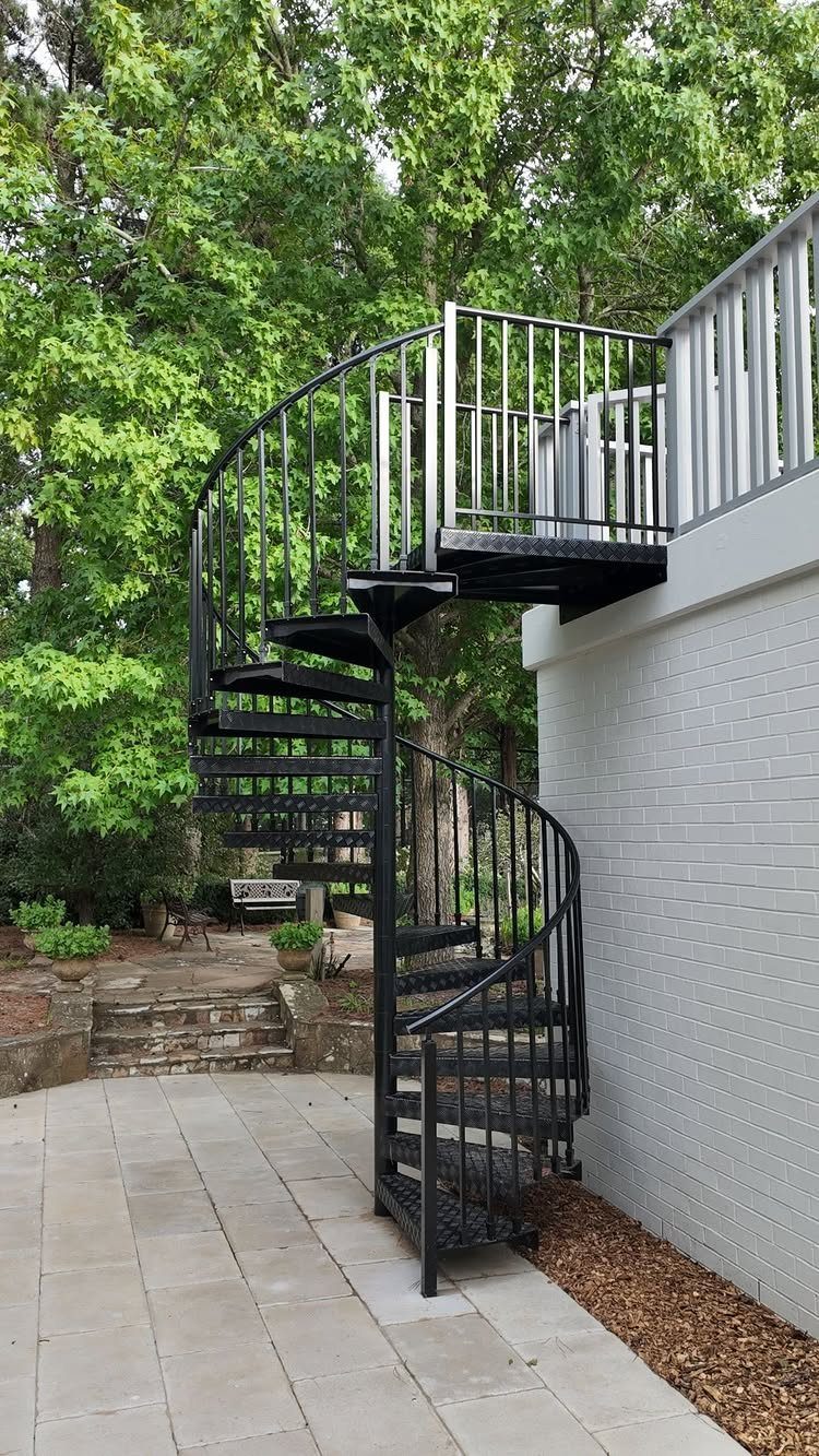 A black metal spiral staircase connecting a stone patio to an elevated grey wooden deck.