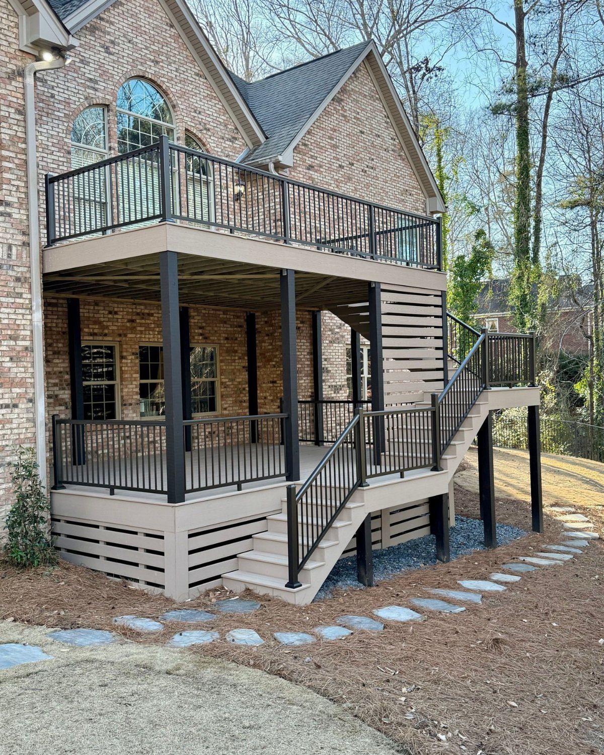 A two-story tan wooden deck with black railings attached to a large brick house.