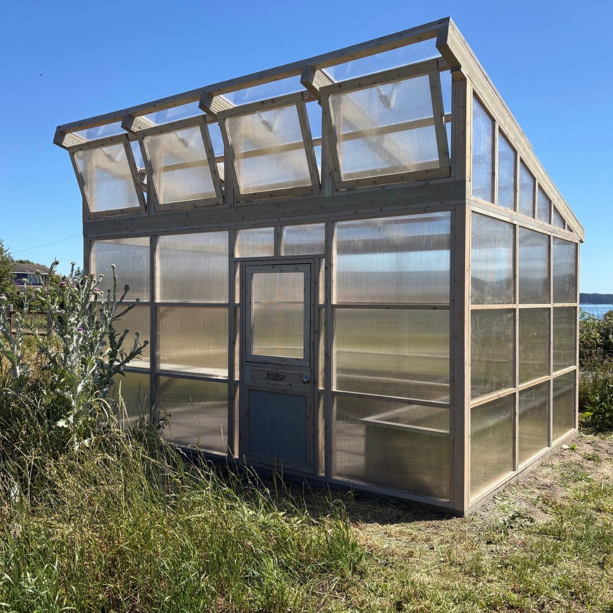 A wooden frame greenhouse with a slanted roof and translucent panels in a grassy field.