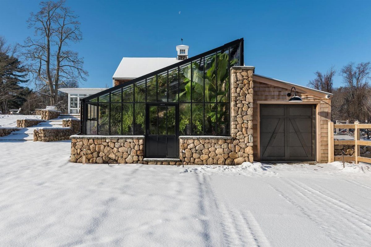 A modern slanted roof greenhouse with black frames and green plants inside, surrounded by snow.