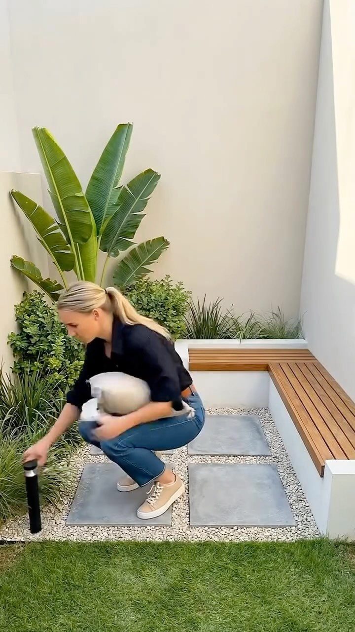 7 Small Yard Garden Ideas for Edibles, Patios and Seating 5 A woman kneels in a modern courtyard garden featuring wooden slat benches and stone pavers.