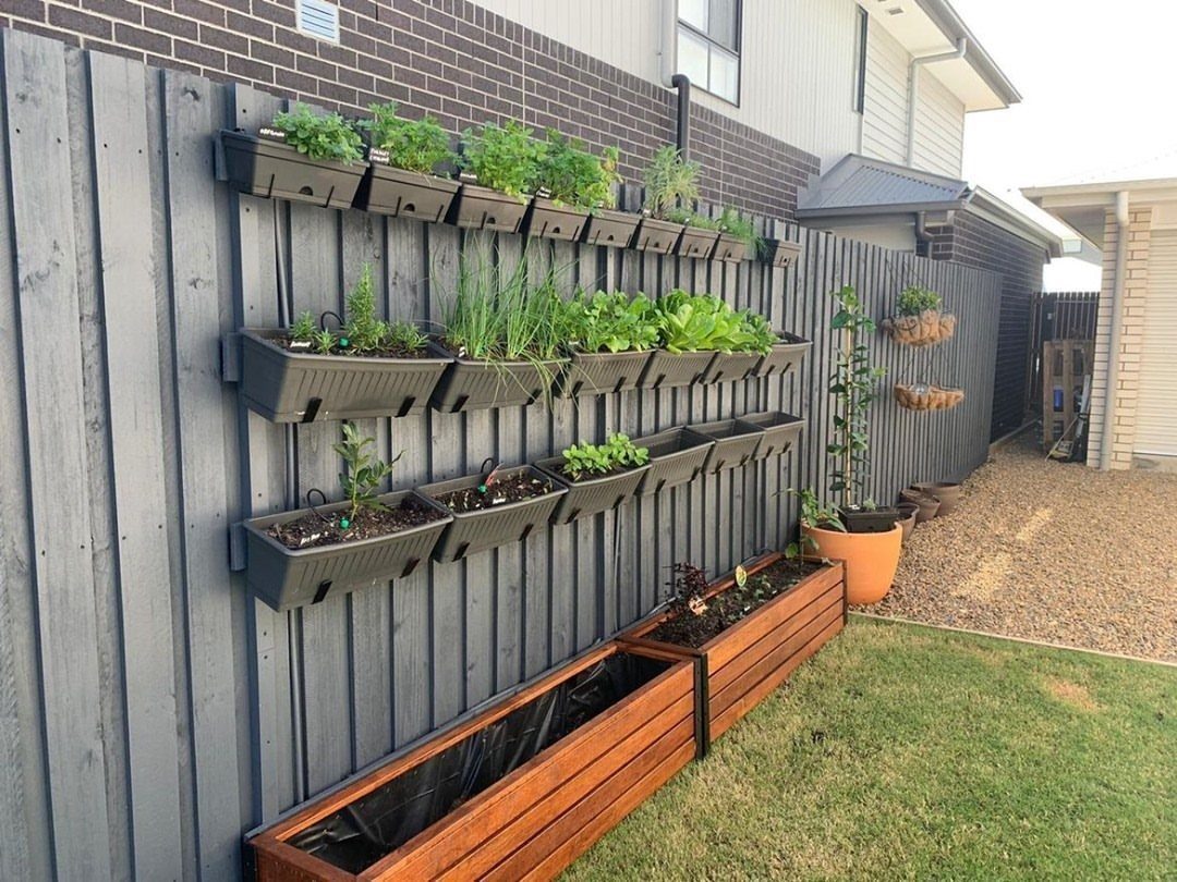 Black rectangular planters filled with green plants mounted vertically on a dark grey wooden fence.