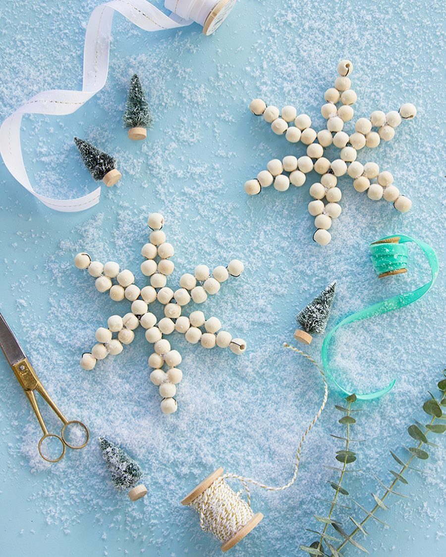 Two snowflake ornaments made of wooden beads resting on artificial snow with ribbons and small trees.