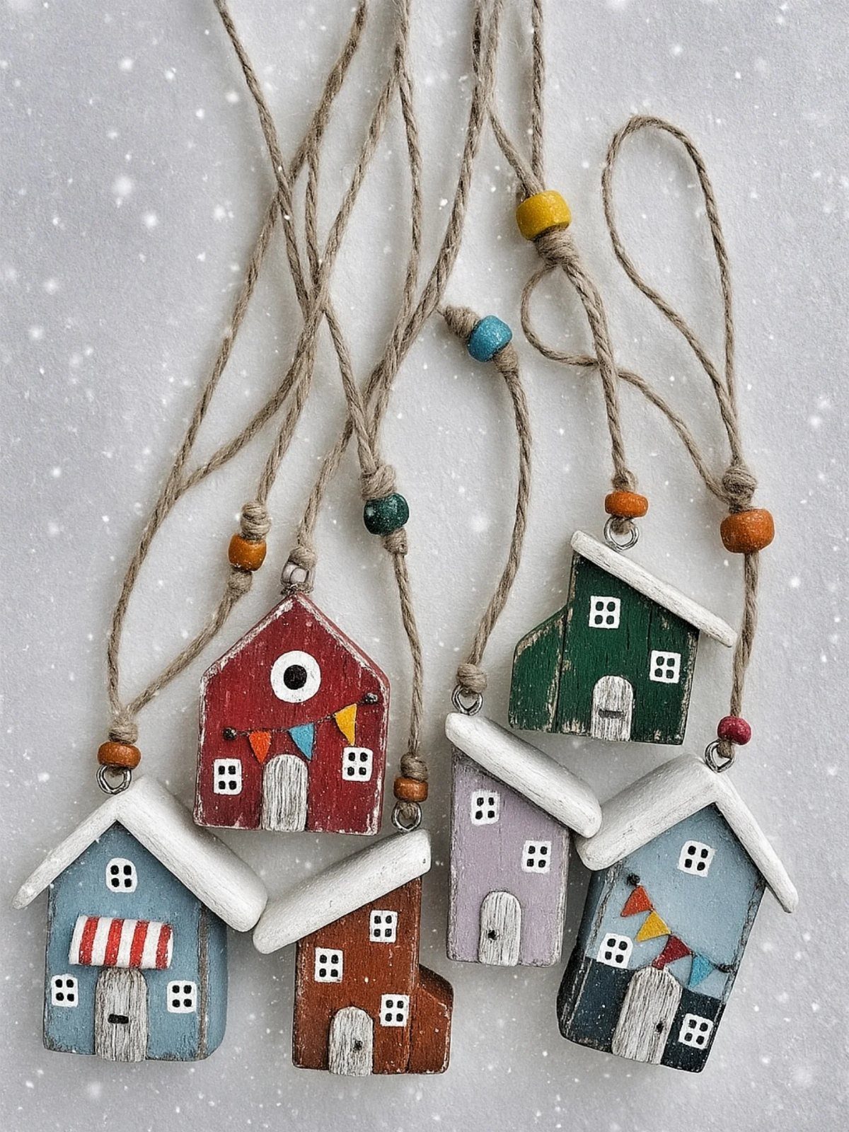 Hand-painted wooden house ornaments in various colors hanging from twine against a snowy background.